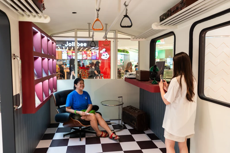 Two guests taking a photograph inside the POKKA coffee train cabin, with vinyl records, upholstered seating, warm lighting and a checkerboard floor.