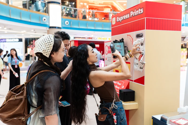 Friends using POKKA’s red-and-cream Express Prints receipt photobooth, with printed photo receipts displayed beside the screen.