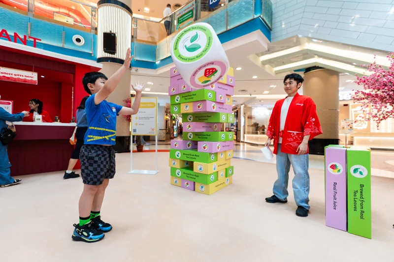 Guest throwing a giant picture die beside green, pink and yellow POKKA Giant Jenga blocks, with a red-jacketed promoter guiding play
