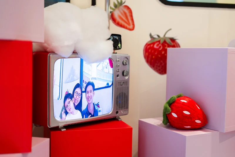 Retro television in the POKKA strawberry cabin showing a family on screen, surrounded by clouds, pastel blocks and strawberry props.
