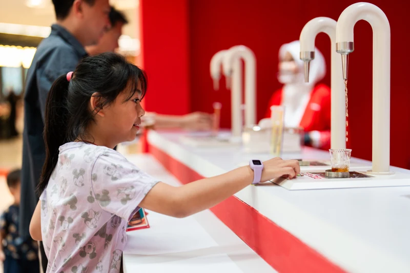 Guest pressing the control at a white curved tap to fill a POKKA sampling cup, with a row of matching taps against the red restaurant counter.