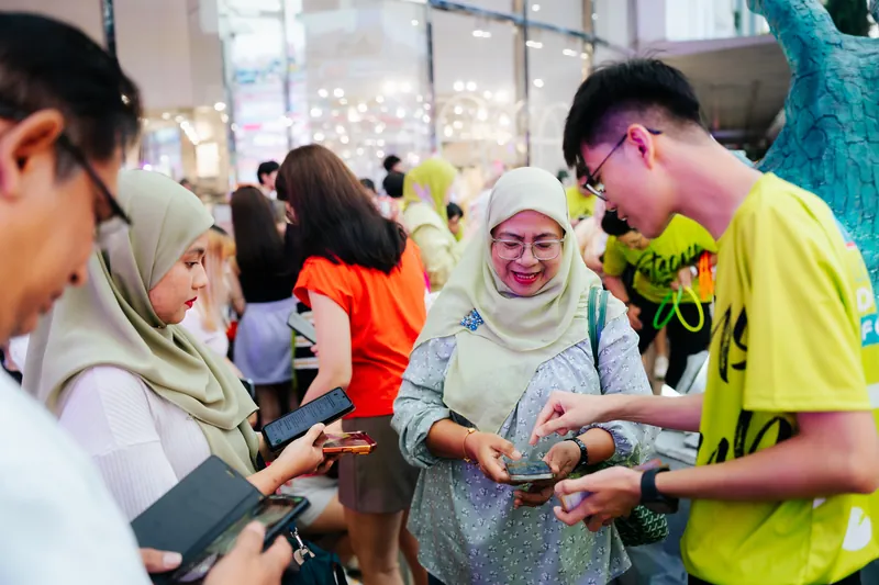 A crew member in a lime campaign T-shirt points at a smiling guest’s phone beside the monster claw as other guests check their own phones, Lot 10, 14 April 2026