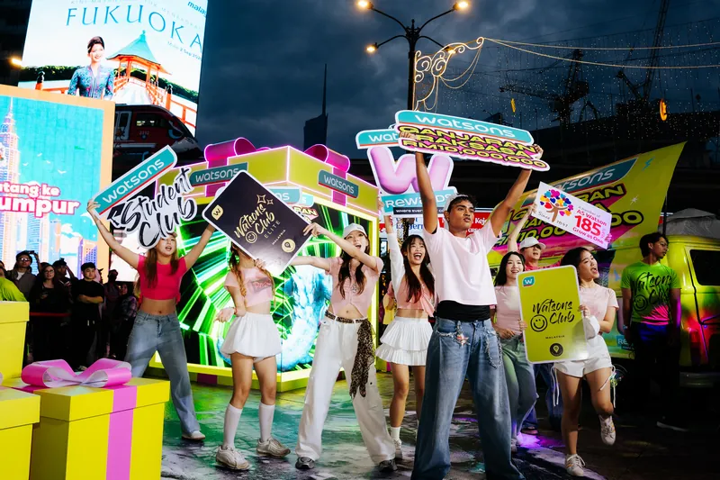 The evening flashmob finale: dancers raise Watsons Club, Student Club and Ganjaran Raksasa placards in front of the glowing LED gift box and the wrapped Kombi van at dusk, Lot 10, 14 April 2026