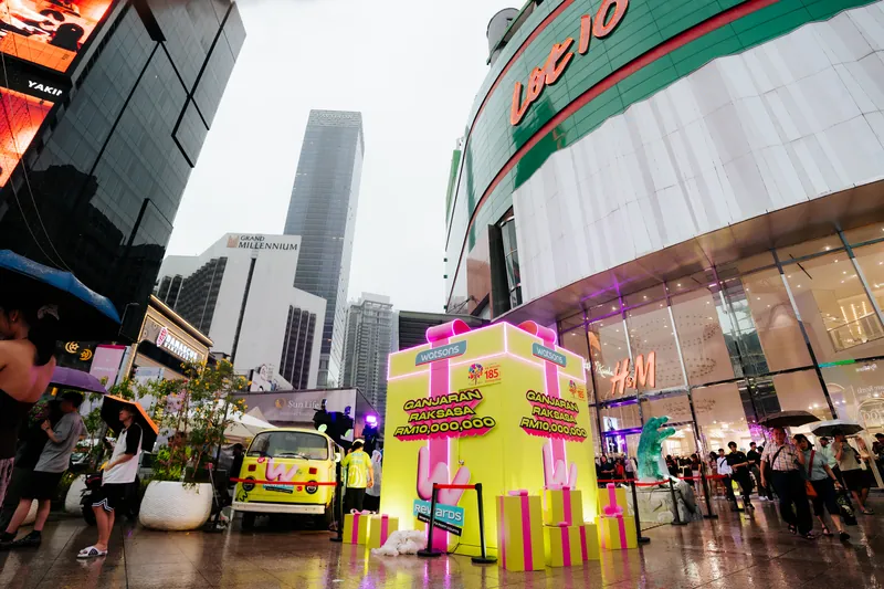 The giant Ganjaran Raksasa gift box at the Lot 10 entrance: a tall yellow box with a pink ribbon and ‘Ganjaran Raksasa RM10,000,000’ lettering, with the wrapped yellow Kombi van beside it, 14 April 2026