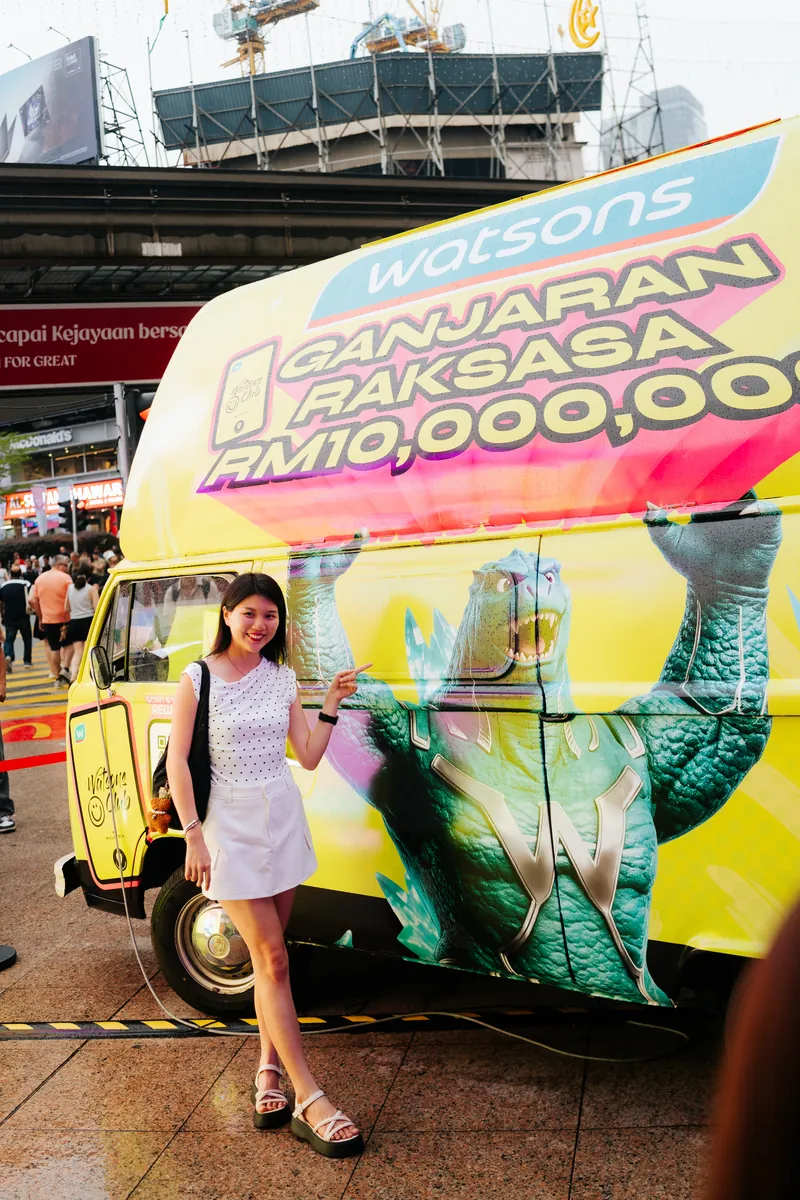A smiling guest in a white polka-dot top and white skirt points at the yellow Kombi van wrapped with a roaring green monster and ‘Ganjaran Raksasa RM10,000,000’ at Lot 10, 14 April 2026