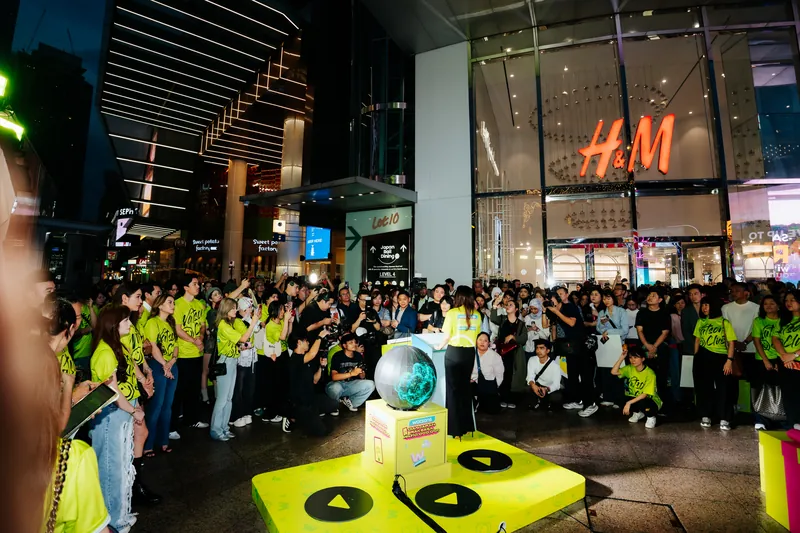 The evening launch ceremony at the Lot 10 entrance: a speaker in a lime T-shirt at a white rostrum beside a glowing LED sphere on a yellow platform, surrounded by photographers and a large crowd outside H&M, 14 April 2026