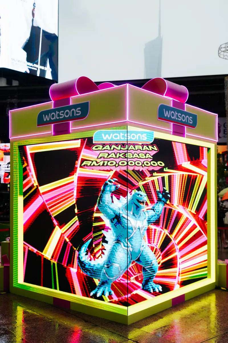 The LED gift box on the Lot 10 plaza playing the Ganjaran Raksasa monster animation: its LED walls show a blue monster against red and yellow neon streaks, topped by a pink bow and Watsons logos, 14 April 2026