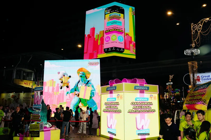 The Lot 10 plaza after dark: the glowing Watsons gift box, a large LED screen showing an animated monster and an LED cube above showing the Watsons app, with guests and crew below, 14 April 2026