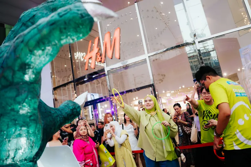 A guest in a green headscarf throws a glowing ring toward the giant monster claw while crew in lime T-shirts and a crowd watch outside the H&M store at Lot 10, 14 April 2026