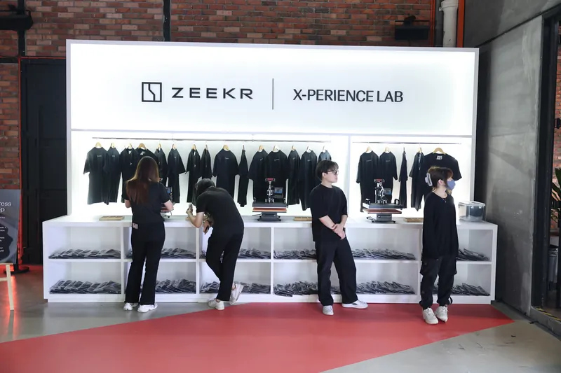 Four crew in black setting up the ZEEKR X-perience Lab heat-press station, T-shirts on the rail above two heat presses, at Odeum, JioSpace