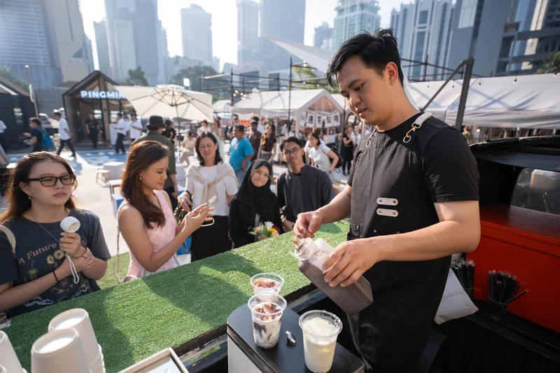 A Galactic Cafe barista pours drinks on the Kombi’s turf-topped counter as guests queue, with the PINGMIN building and city towers behind.