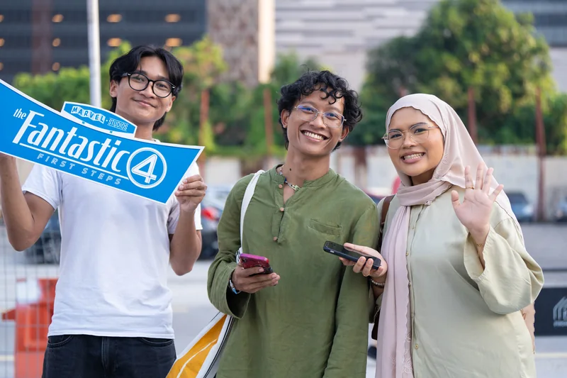 A crew member in a white tee holds up a Fantastic Four: First Steps placard beside two smiling guests with their phones.