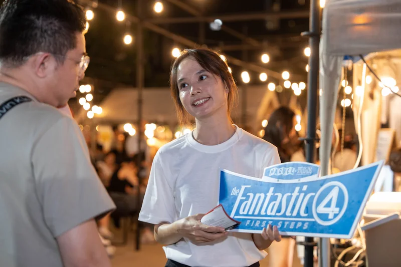 A crew member in a white tee holds a Fantastic Four: First Steps placard and a card as she talks to a guest, under string lights in the night market.