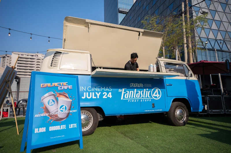 The Galactic Cafe: Baito’s vintage Kombi in the film’s blue livery, serving hatch open and a barista inside, beside an A-frame sign reading ‘Redeem your coffee now!’ with Black, Chocolate and Latte.