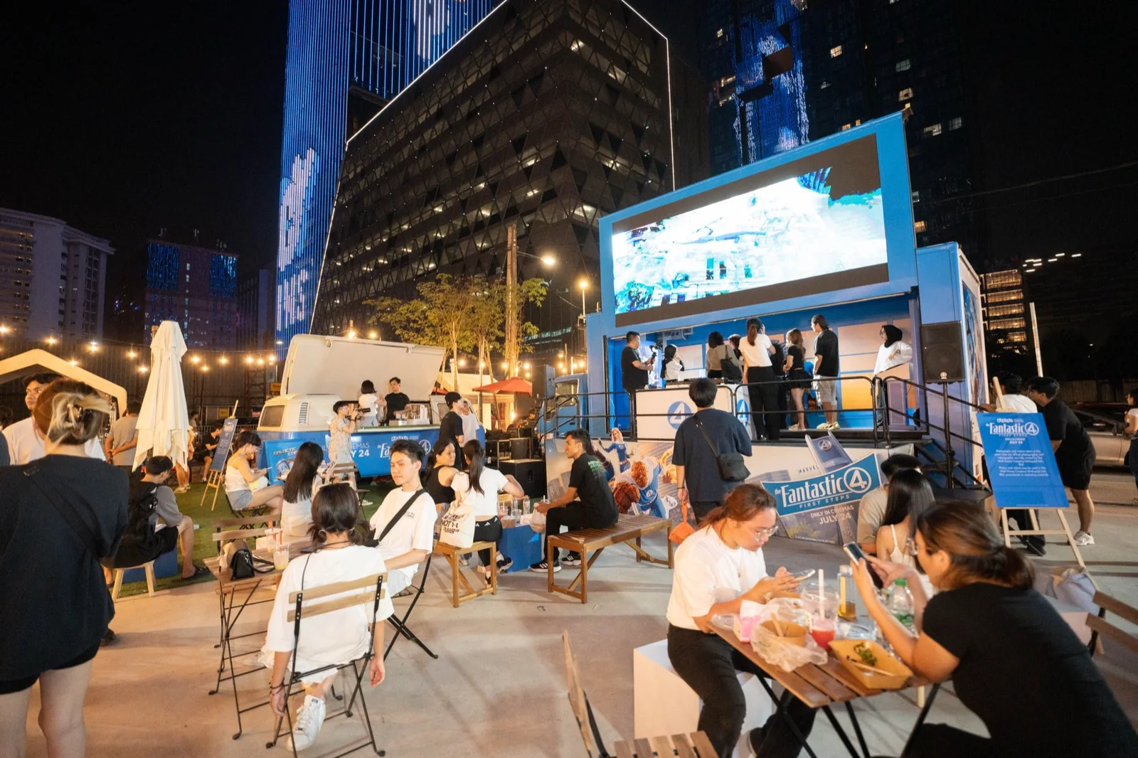 The wrapped stage truck at Pingmin Hub, Kuala Lumpur, after dark: film footage on its LED screen, people on its stage, the Galactic Cafe Kombi behind, and guests at tables, benches and cube seats in front.