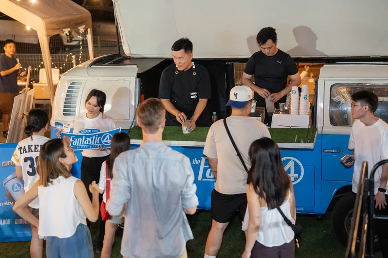 Guests queue at the Galactic Cafe Kombi after dark as two baristas pour drinks, and a crew member in a white tee holds a Fantastic Four: First Steps placard.