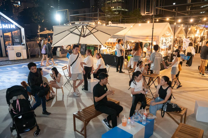 Guests at benches and cube seats on the pop-up’s blue floor at night, with Pingmin Hub’s market tents, parasols and festoon lights behind.