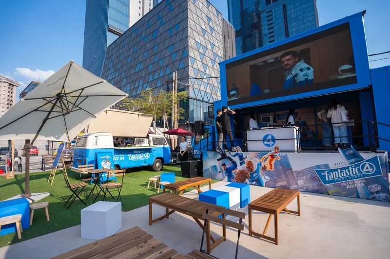 The pop-up in late-afternoon light: the blue Galactic Cafe Kombi and the stage truck behind turf, parasols, wooden benches and cube seats, with a Fantastic Four: First Steps panel reading ‘Only in cinemas July 24’.