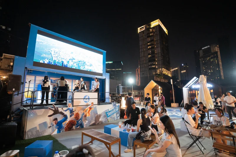 The wrapped stage truck at Pingmin Hub after dark: film footage on its LED screen, people on its stage, a Fantastic Four: First Steps character panel along the front, and guests at wooden benches and cube seats.
