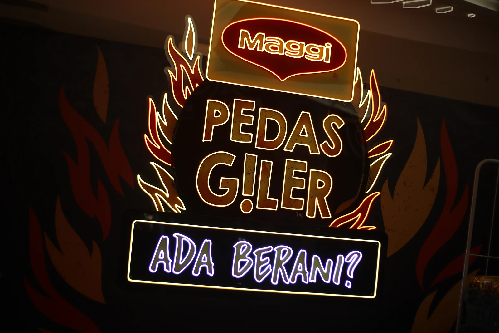 The MAGGI Pedas Giler neon sign with ‘Ada Berani?’ lit in purple beneath it, on a black wall painted with flames.