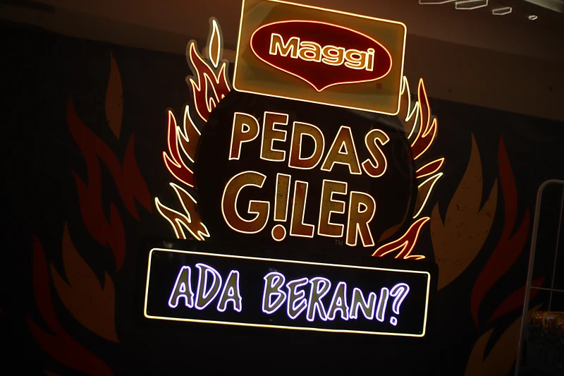 The MAGGI Pedas Giler neon sign with ‘Ada Berani?’ lit in purple beneath it, on a black wall painted with flames.