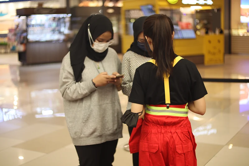 A crew member in red coveralls and yellow braces stands with two visitors as they use their phones near the registration point.