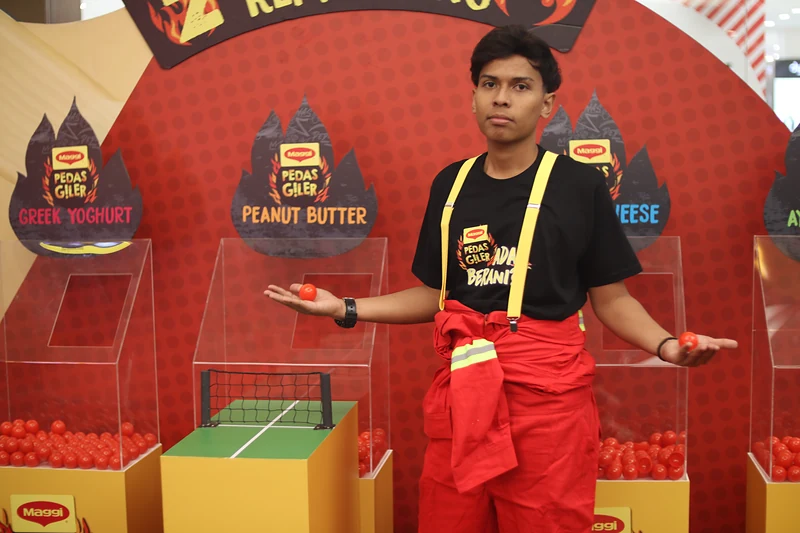 A crew member in the ‘Ada Berani?’ tee, yellow braces and red coveralls holds out two red balls in front of the Remix Pong bins.