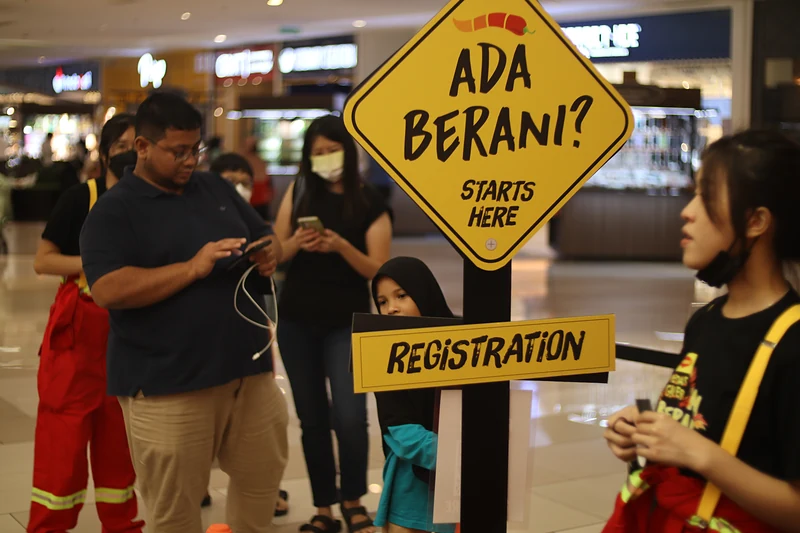 Shoppers check their phones in the queue at the ‘Ada Berani? Starts here’ registration sign, with crew in red coveralls beside them.