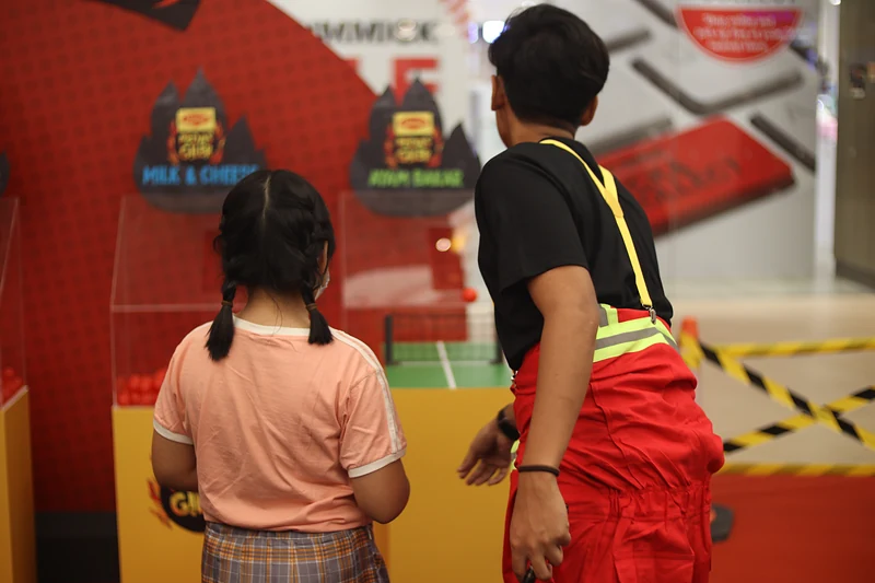 A young player, seen from behind, lines up a throw at Remix Pong beside a crew member in red coveralls.