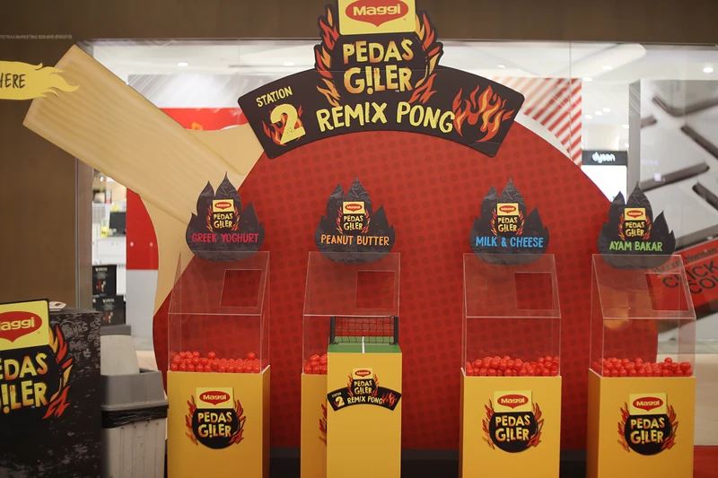 Station 2, Remix Pong: four clear bins labelled Greek Yoghurt, Peanut Butter, Milk & Cheese and Ayam Bakar on yellow MAGGI Pedas Giler plinths, with a mini net and trays of red balls.