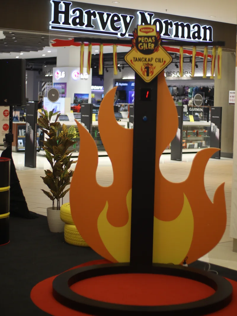 Station 1, Tangkap Cili: chilli-printed sticks hang from an arm above a black pole with a digital counter, set against tall orange flame cut-outs on a red ring.