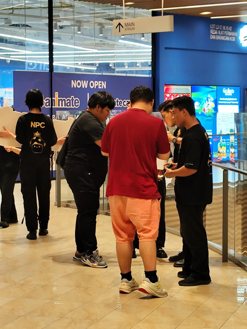 Crew members, one in an NPC tee holding a cut-out, stand with shoppers looking at their phones and papers on a mall concourse.