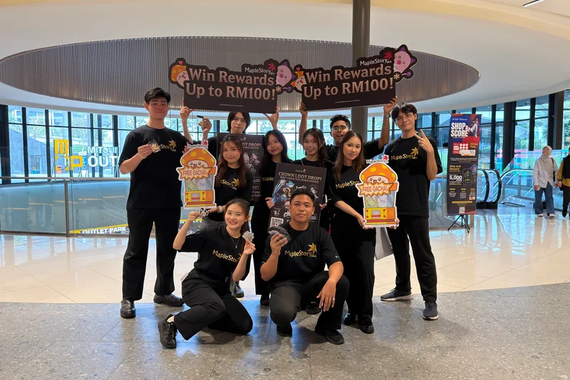 Ten crew in black MapleStorySEA tees pose in a LaLaport atrium with two ‘Win Rewards Up to RM100!’ signs, two ‘FREE GACHA!’ cut-outs and a Crown Loot Drop poster.