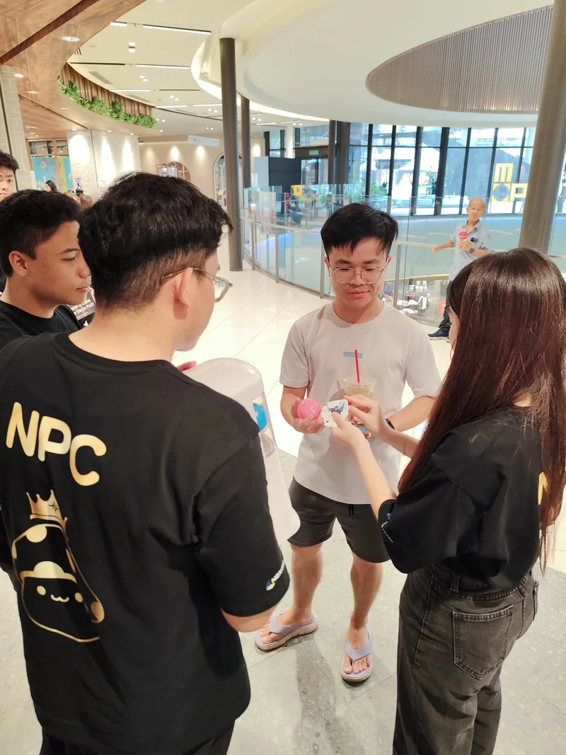 A shopper holds an opened pink capsule and a slip of paper, facing a crew member in an NPC tee and another crew member in a MapleStorySEA tee.