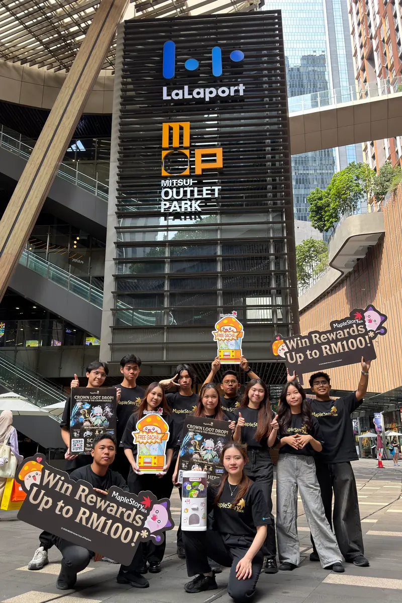 Eleven crew in MapleStorySEA tees with signs, posters, cut-outs and the capsule machine below the LaLaport and Mitsui Outlet Park tower at the mall entrance.