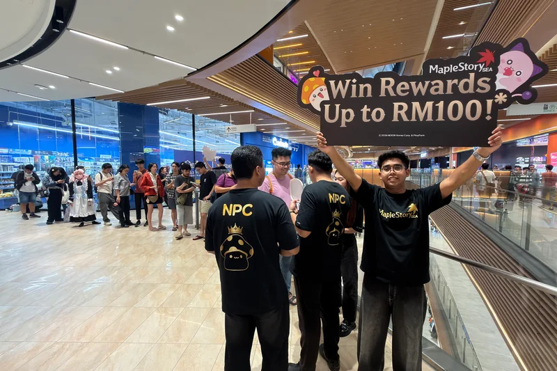 Two crew members seen from behind in black tees printed with ‘NPC’ and a crowned mushroom, beside a crew member holding up a MapleStorySEA ‘Win Rewards Up to RM100!’ sign, while shoppers queue with their phones in a LaLaport atrium.