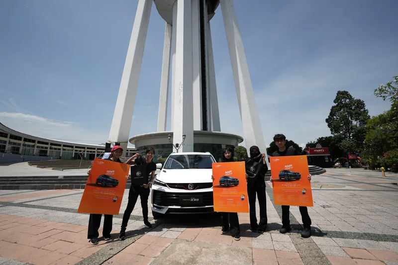Crew in black hold orange Roar of Amazement boards around a white X50 at the foot of Alor Setar Tower