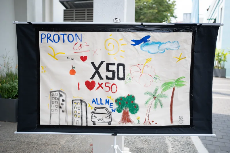 A painted canvas reading ‘PROTON’, ‘X50’ and ‘I ♥ X50’, with palm trees, buildings and a car, on a stand outside Central Market, Kuala Lumpur
