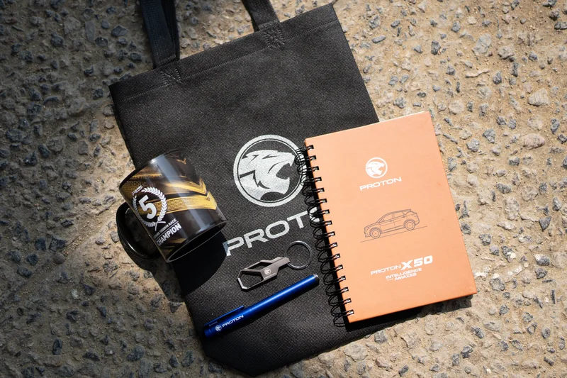 PROTON premiums laid out on the ground: a black tote bag, a mug, a keychain, a pen and an orange X50 notebook