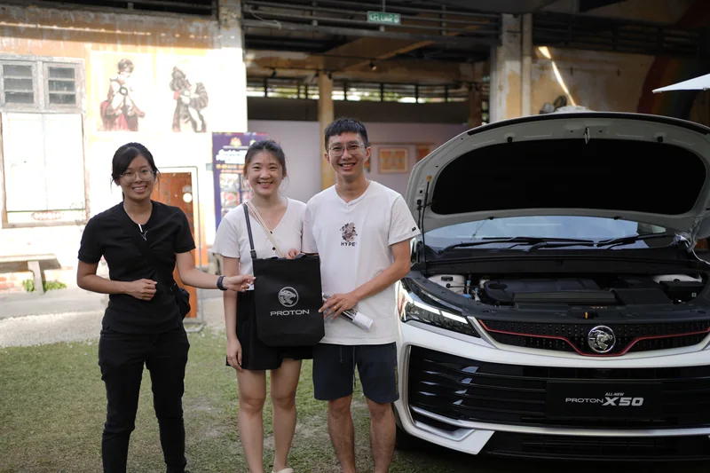 A crew member in black stands with two visitors holding a PROTON tote bag beside a white X50 with its bonnet open at Hin Bus Depot, George Town