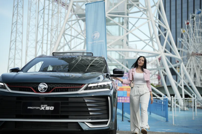 A woman in pink walks past a dark X50 in front of the white lattice tower at i-City, Shah Alam