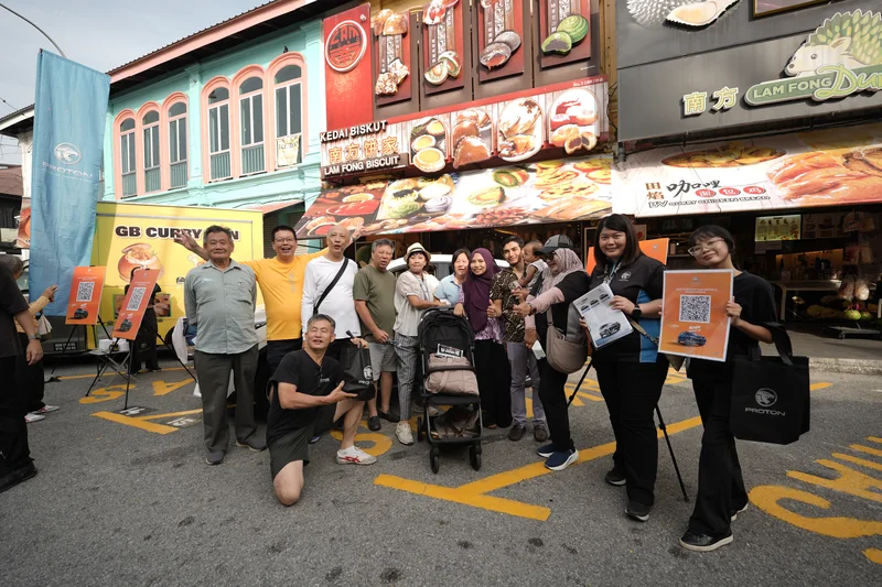 Two crew members in black, one holding an orange sign-up board, with a crowd of cheering visitors outside the shophouses of Ipoh old town