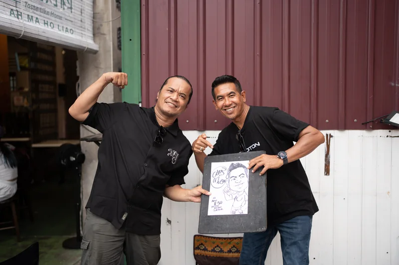 A caricature artist and a man in a PROTON shirt pose on Jonker Street with a finished sketch marked ‘All New X-50 PROTON’