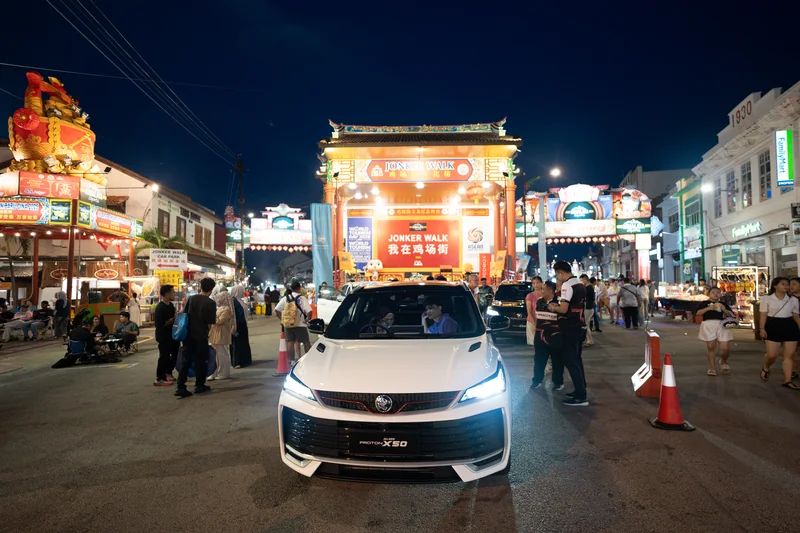 A white X50 with its headlights on in front of the lit Jonker Walk gateway on Jonker Street, Melaka, at dusk, with crowds on both sides