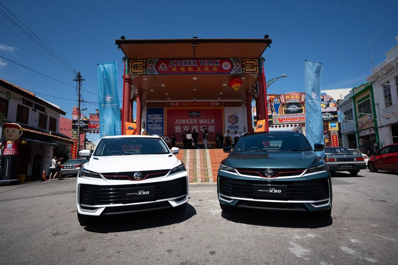 A white and a dark X50 parked side by side in front of the red Jonker Walk stage under a blue sky, Melaka