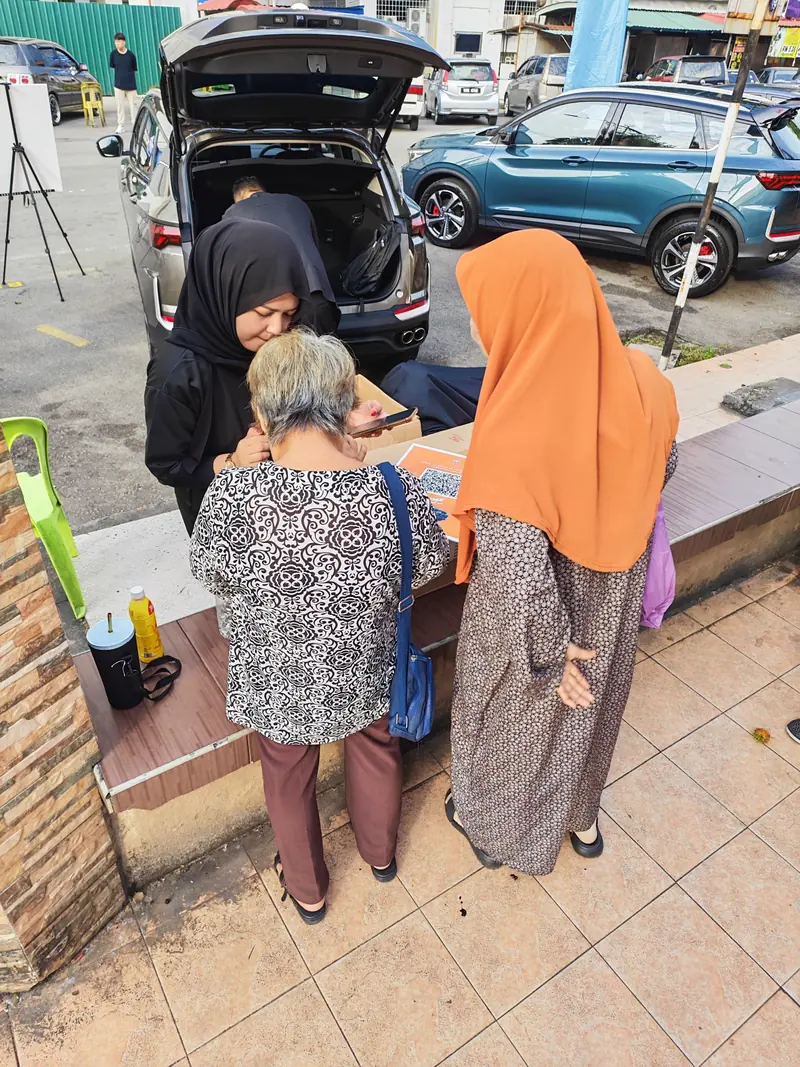 A crew member and two visitors look at a phone together beside an X50 with its boot open in Kota Bharu