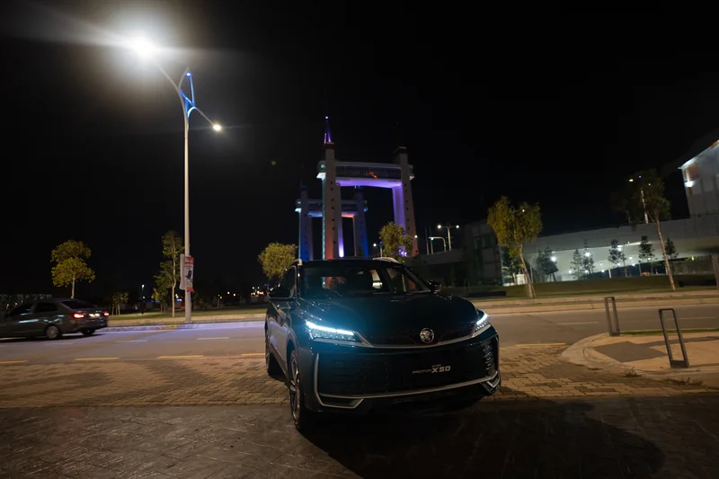 A dark X50 with its lights on under a street lamp at night, with the lit Kuala Terengganu drawbridge behind