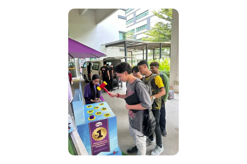 A student swings the red mallet at the blue Ribena whack-a-mole unit, its purple blackcurrant ‘moles’ set in yellow rings, as friends queue behind him and a Baito crew member stands beside it at a roving stop