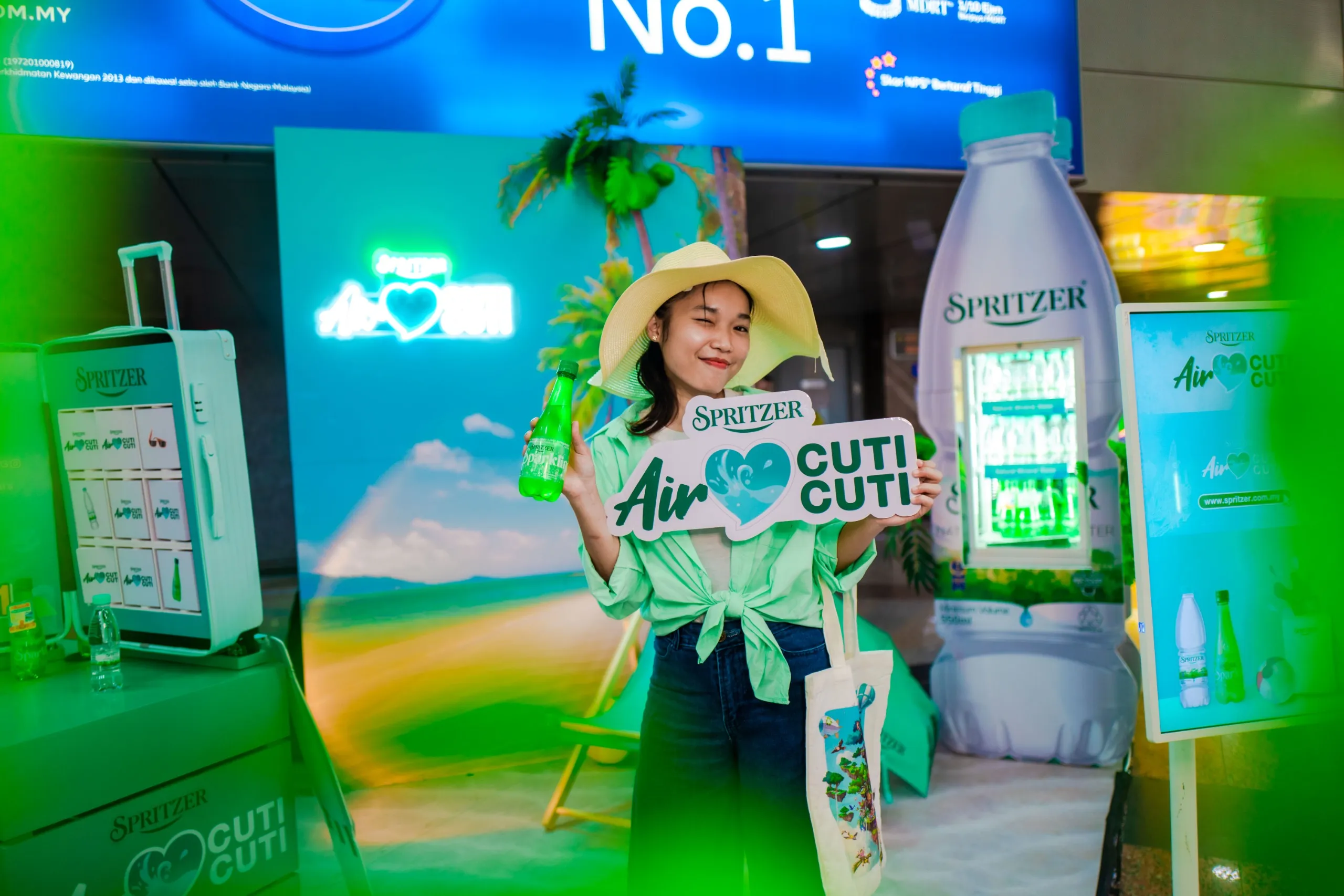 A woman in a sun hat holding a Spritzer Sparkling bottle and an Air Cuti Cuti sign at a Spritzer Air Cuti Cuti booth.